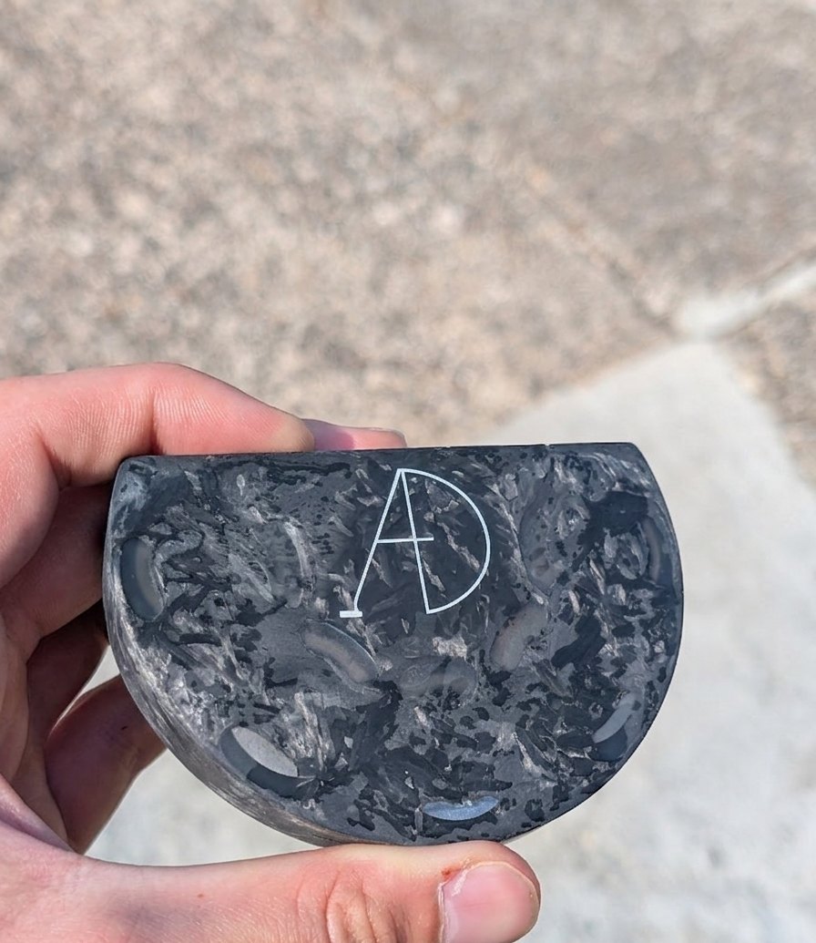 The current prototype putter head held in hand, its carbon fibre surface marbled and engraved with the AD mark
